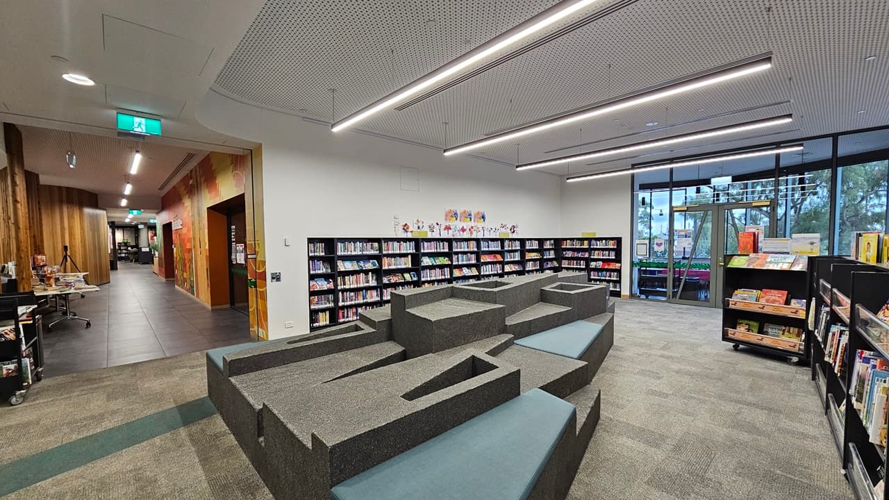On the left, there is a hallway with wooden walls and a green exit sign above the door. In the middle, there is a grey, tiered seating area with blue cushions and integrated steps. On the right, there are bookshelves filled with books and a large window with a view of trees and buildings outside. The floor is covered in grey carpet tiles with a green stripe, and the ceiling is white with a perforated design and linear recessed lighting.