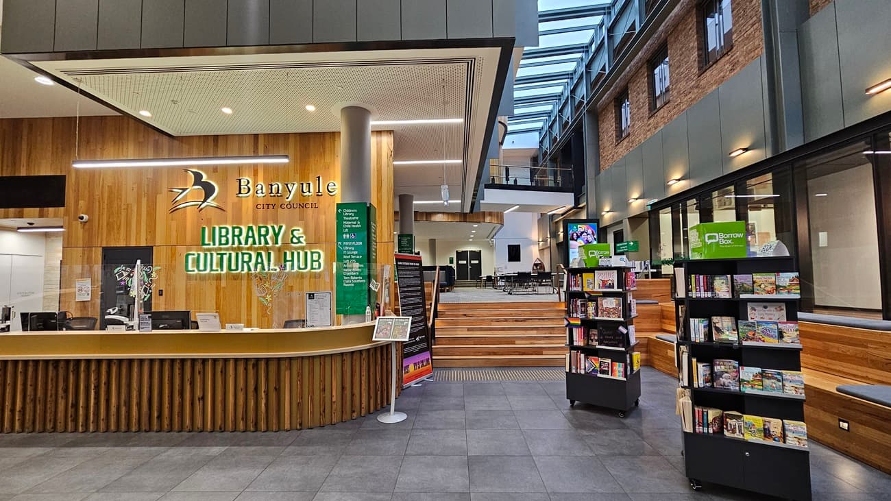 On the left, there is a wooden reception counter with the text "Banyule City Council Library & Cultural Hub" on the wall behind it, along with a green information board. In the middle, a set of wooden steps leads to an open seating area with tables and chairs. On the right, there are bookshelves filled with books and a green sign. The floor is covered with large grey tiles, and the ceiling is white with recessed lighting and a skylight above the seating area.