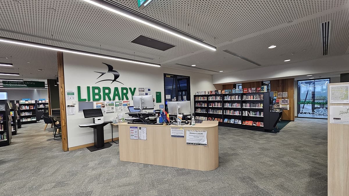 On the left, there is a white wall with the word "LIBRARY" in green and a black and white bird logo above a computer desk with monitors and office supplies. In the middle, there is a curved wooden reception desk with notices and a computer. On the right, there are bookshelves filled with books and a large window with a view of trees. The floor is covered in grey carpet tiles, and the ceiling is white with perforated panels and recessed lighting.