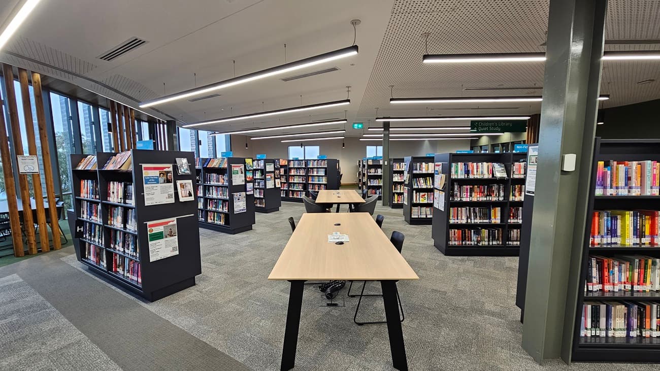 On the left, there are black bookshelves filled with books and a wall with vertical wooden slats and large windows. In the middle, there are long wooden tables with black chairs and more bookshelves behind them. On the right, there are additional black bookshelves filled with books and a grey column. The floor is covered in grey carpet tiles, and the ceiling is white with perforated panels and recessed linear lighting.