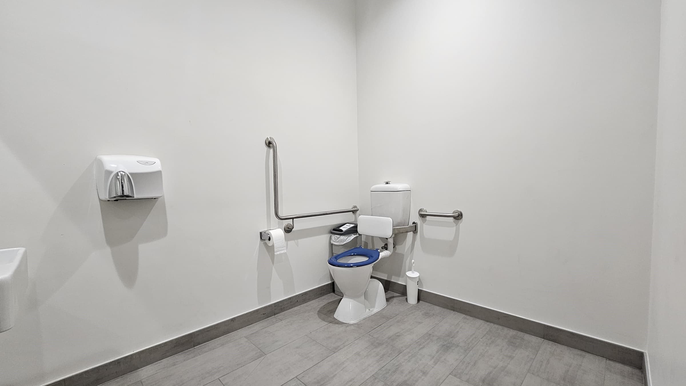On the left, there is a white wall-mounted soap dispenser. In the middle, there is a white toilet with a blue seat, equipped with stainless steel grab bars on both sides and a white toilet paper holder. On the right, there is a white wall with no fixtures. The floor is covered in light grey tiles, and the ceiling is white with recessed lighting.