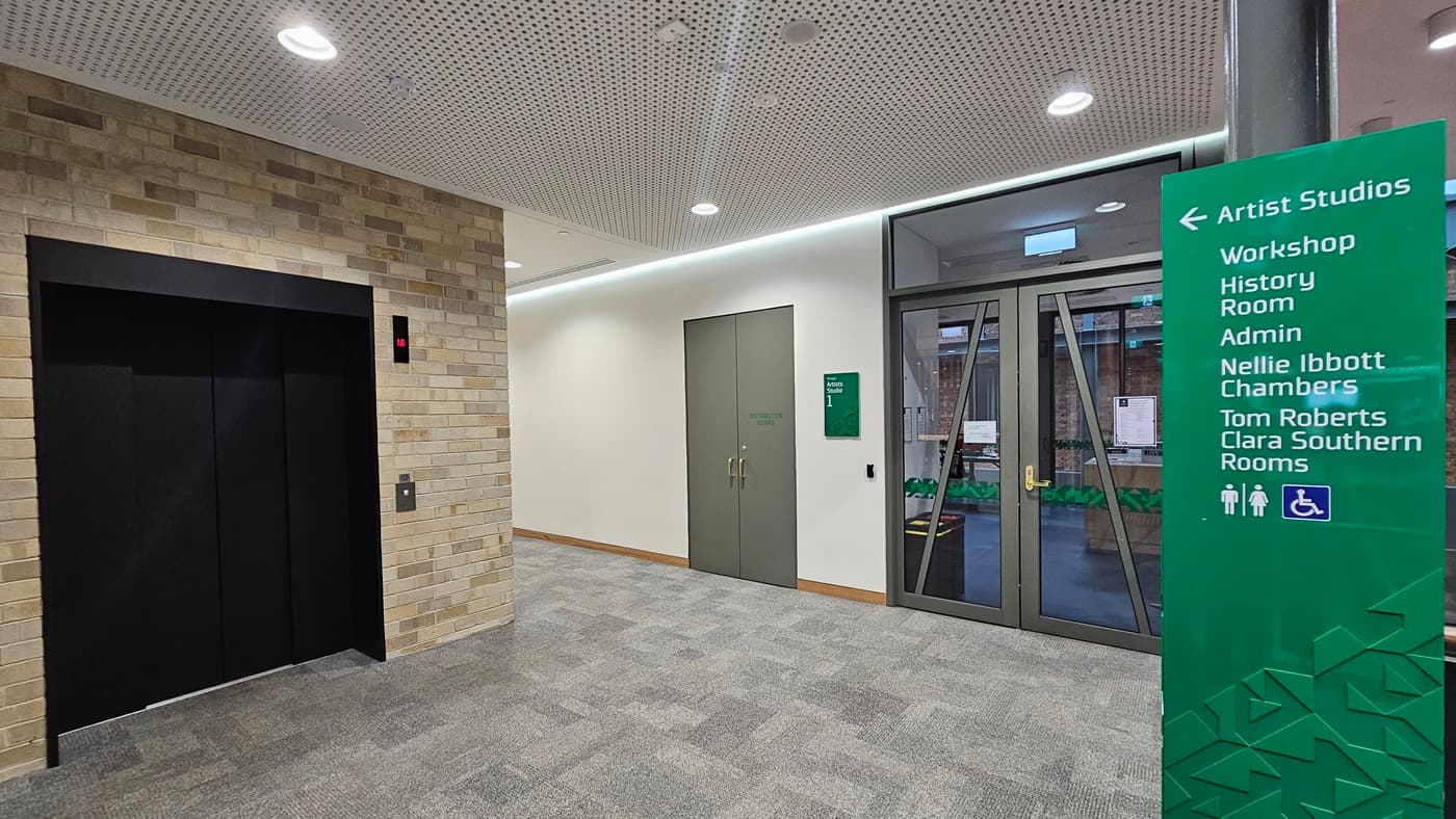 On the left, there is a black lift with a control panel and a red indicator light, set in a wall of light-coloured bricks. In the middle, there are two grey double doors with a green sign next to them. On the right, there is a glass door with a view of an outdoor area and a green sign listing room names and directions. The floor is covered in grey carpet tiles, and the ceiling is white with perforated panels and recessed lighting.