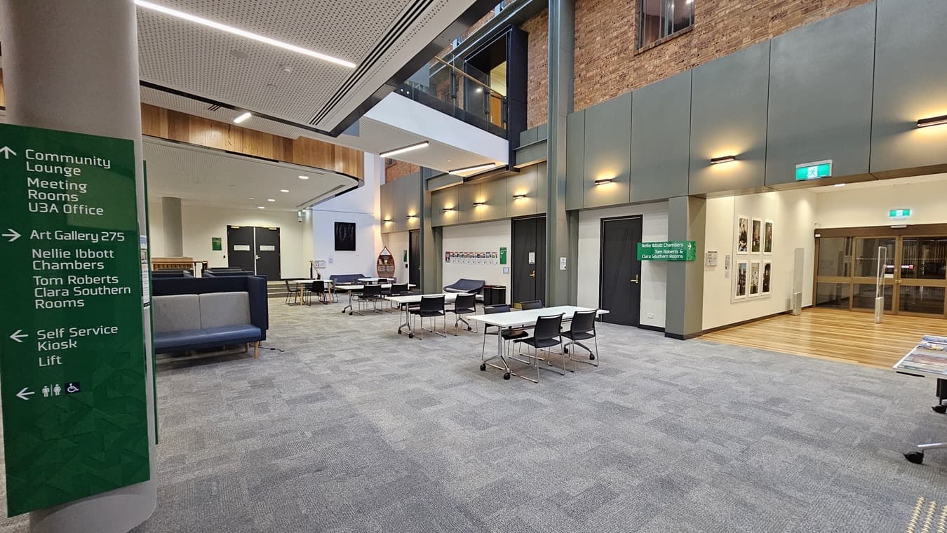 On the left, there is a green sign listing various rooms and facilities. In the middle, there are tables and chairs arranged in an open lounge area with a grey carpeted floor. In the background, there are black double doors and a staircase leading to an upper level with glass railings. On the right, there is a brick wall with framed pictures, a green exit sign, and a wooden reception counter with brochures. The ceiling is white with recessed lighting and a perforated panel design.