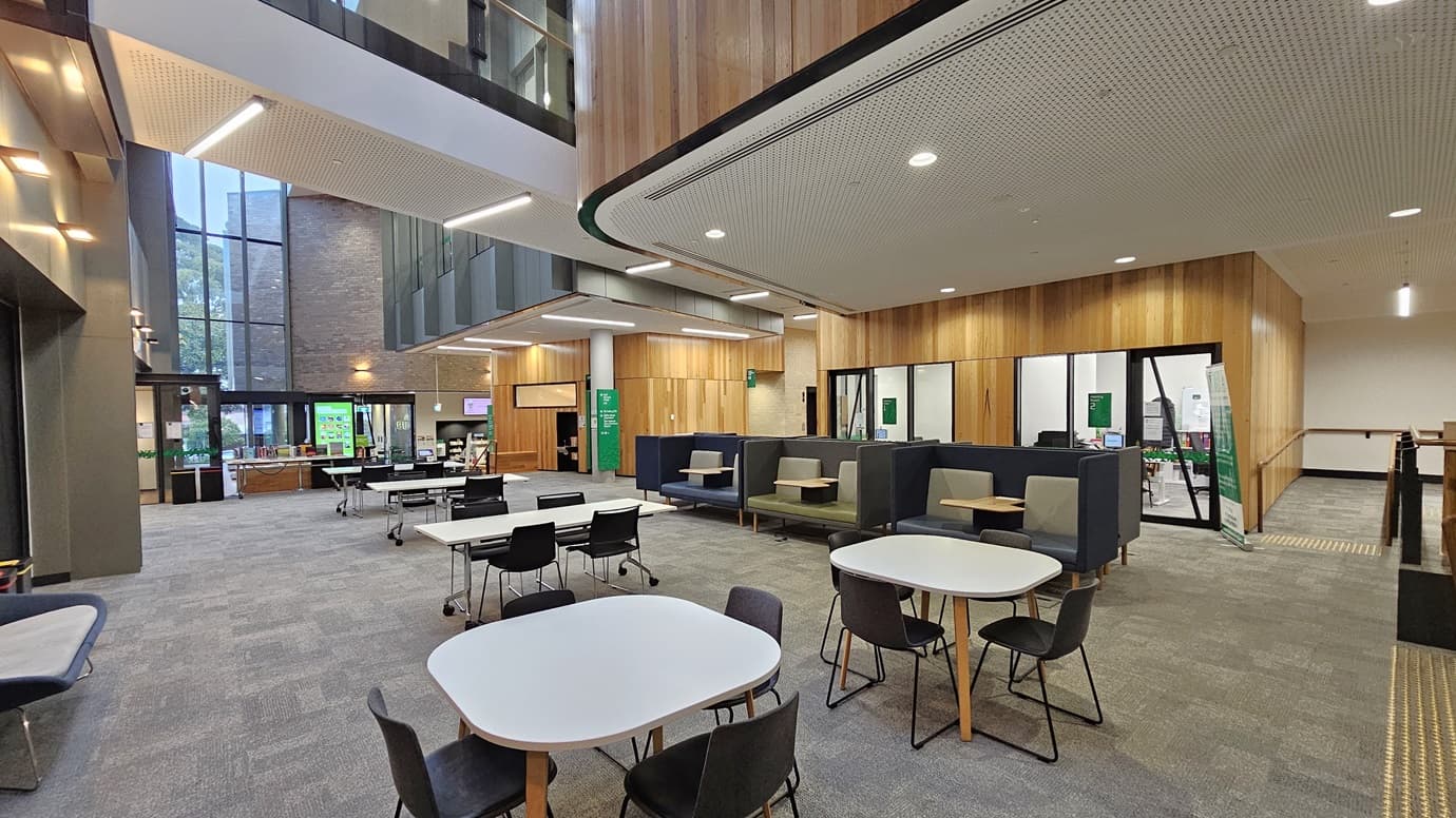 On the left, there are tall windows and a seating area with benches and tables. In the middle, there are several tables and chairs arranged in an open lounge area with grey carpeted flooring. In the background, there is a reception desk with a green sign and a brick wall. On the right, there are enclosed office spaces with glass walls and wooden panelling. The ceiling is white with recessed lighting and perforated panels.