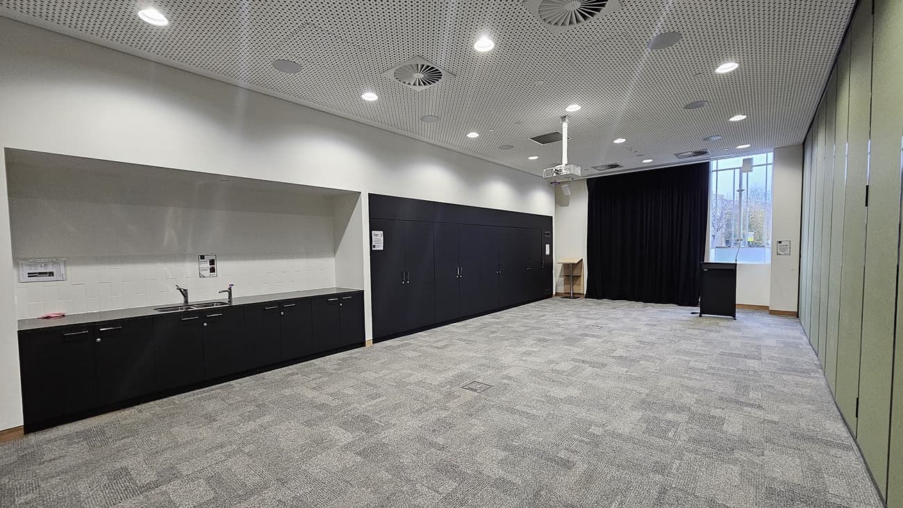 On the left, there is a long black counter with a sink, cabinets, and a white backsplash. In the middle, there is an open space with grey carpet tiles. In the background, there is a black curtain, a podium, and a window with a view of trees. On the right, there is a green partition wall. The ceiling is white with recessed lighting, circular air vents, and perforated panels.