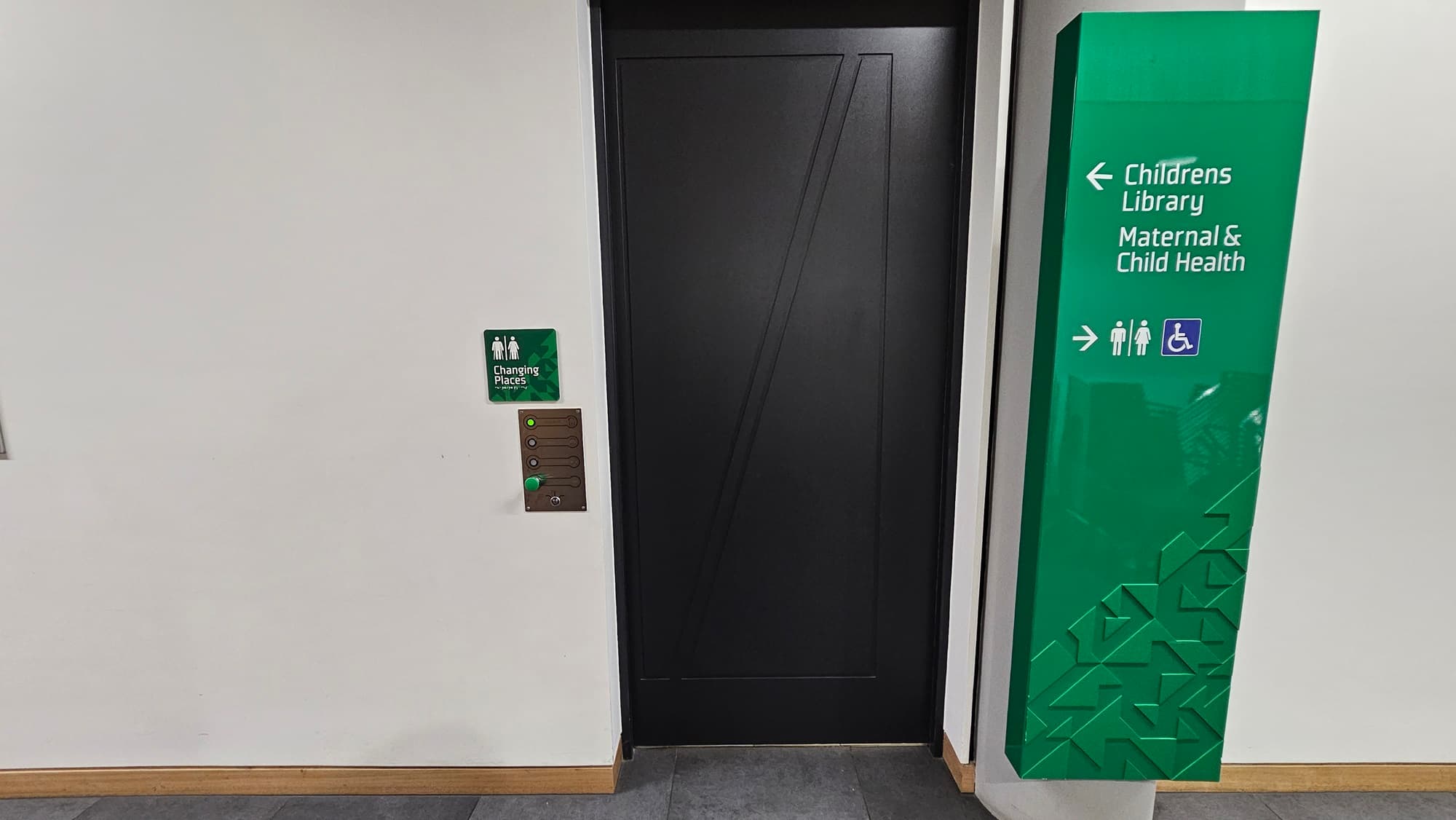 On the left, there is a white wall with a small green sign indicating "Change Project Room" and an intercom with buttons. In the middle, there is a black door with a geometric pattern. On the right, there is a large green sign with white text pointing to the "Children's Library" and "Maternal & Child Health" with icons for accessibility. The floor is covered with grey tiles, and the baseboard is wooden.