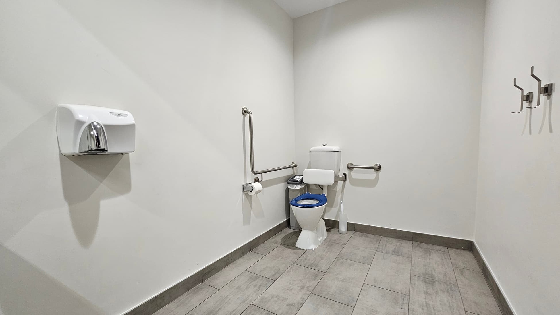 On the left, there is a white wall-mounted paper towel dispenser. In the middle, there is a white toilet with a blue seat, flanked by metal grab bars and a toilet paper holder. On the right, there are three metal coat hooks attached to the wall. The floor is covered with large grey tiles, and the ceiling is white with a slight slope.