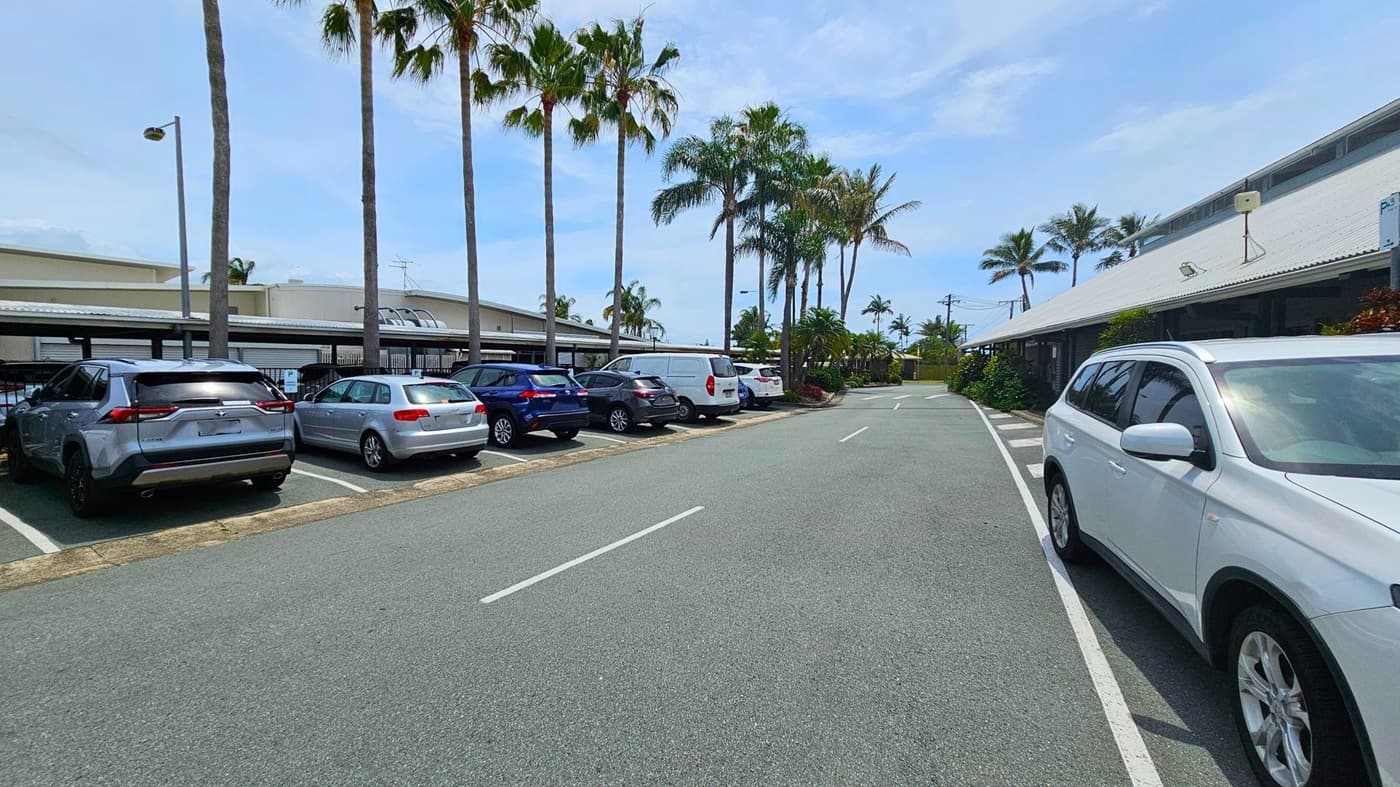 A car park with several vehicles on the left, including a silver SUV and a blue car. Tall palm trees line the area, providing shade. On the right, a white SUV is parked near a building with a sloped roof. The road in the centre is clear, with white lines marking the lanes. The background features more palm trees and a few low-rise buildings.