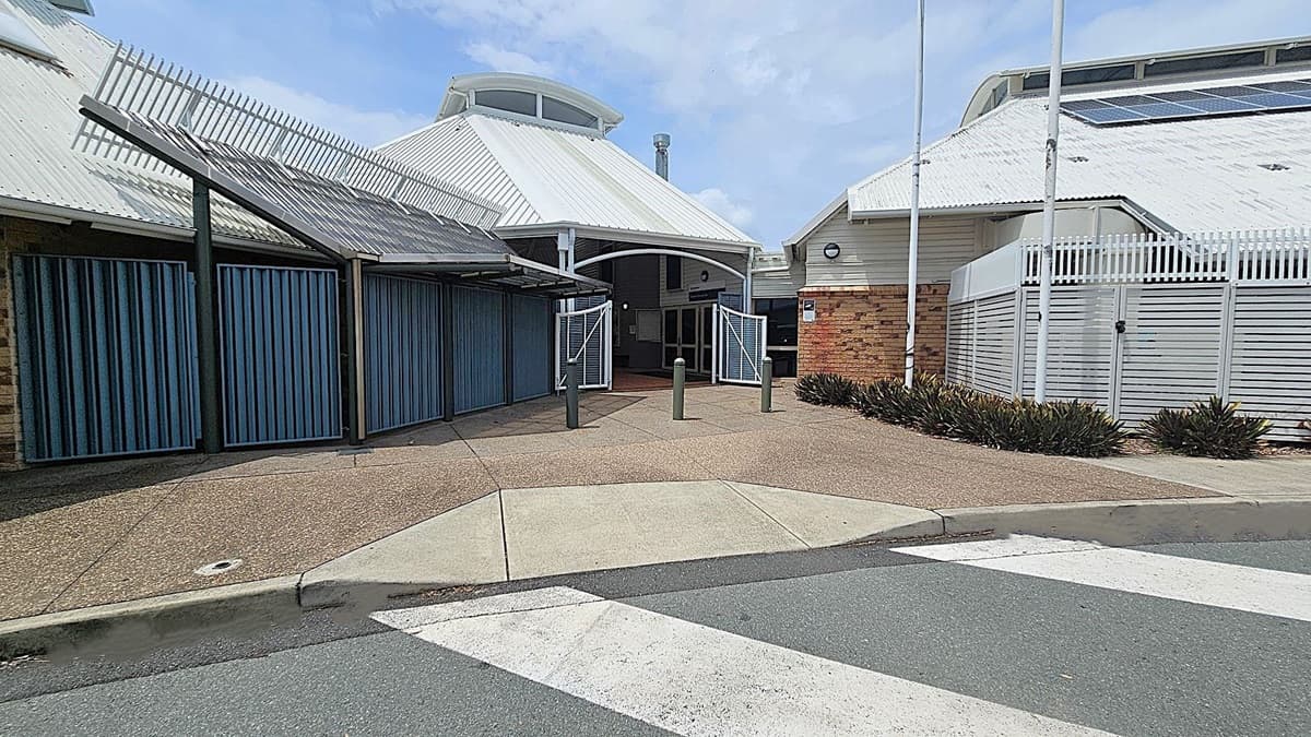 A building with a white corrugated metal roof and solar panels. The entrance features blue double doors and is flanked by brick and metal fencing. The foreground has a textured pavement with a small green section and a zebra crossing. There are bollards near the entrance and some low shrubs to the right. The building has a modern design with angular lines and a mix of materials.