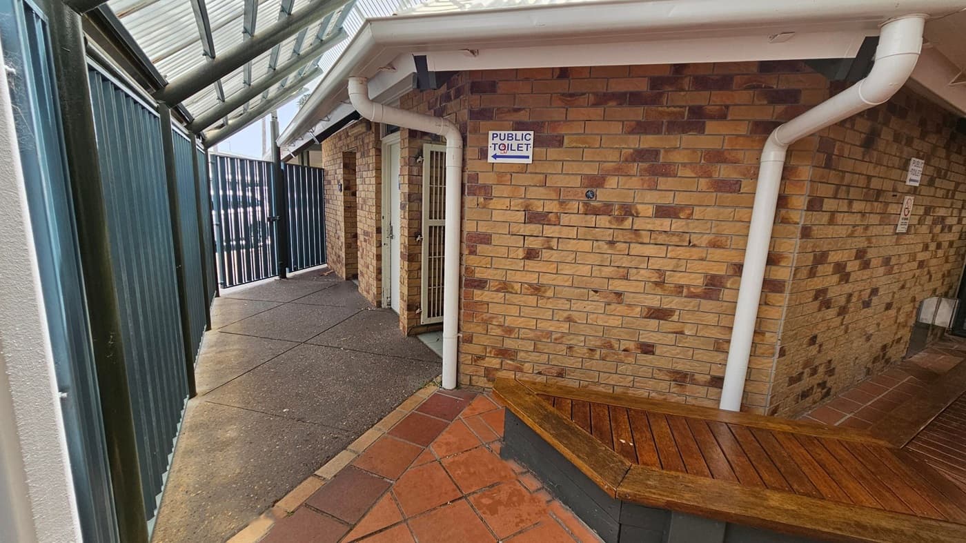 A narrow walkway with a corrugated metal roof on the left, leading to a gate. The path is paved with concrete and tiles. On the right, a brick building with a sign indicating a public toilet. A wooden bench is attached to the building. White downpipes run along the building's corners. The floor is a mix of red and brown tiles.