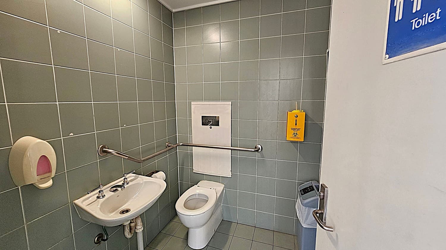 A public toilet with green tiled walls and a green tiled floor. On the left, there's a small white sink with a soap dispenser above it and a metal handrail beside it. In the centre, a white toilet with a handrail on the wall. Above the toilet is a white panel, and to the right, a yellow sanitary disposal box. On the right, a partially open door with a blue sign indicating "Toilet" and a waste bin nearby.