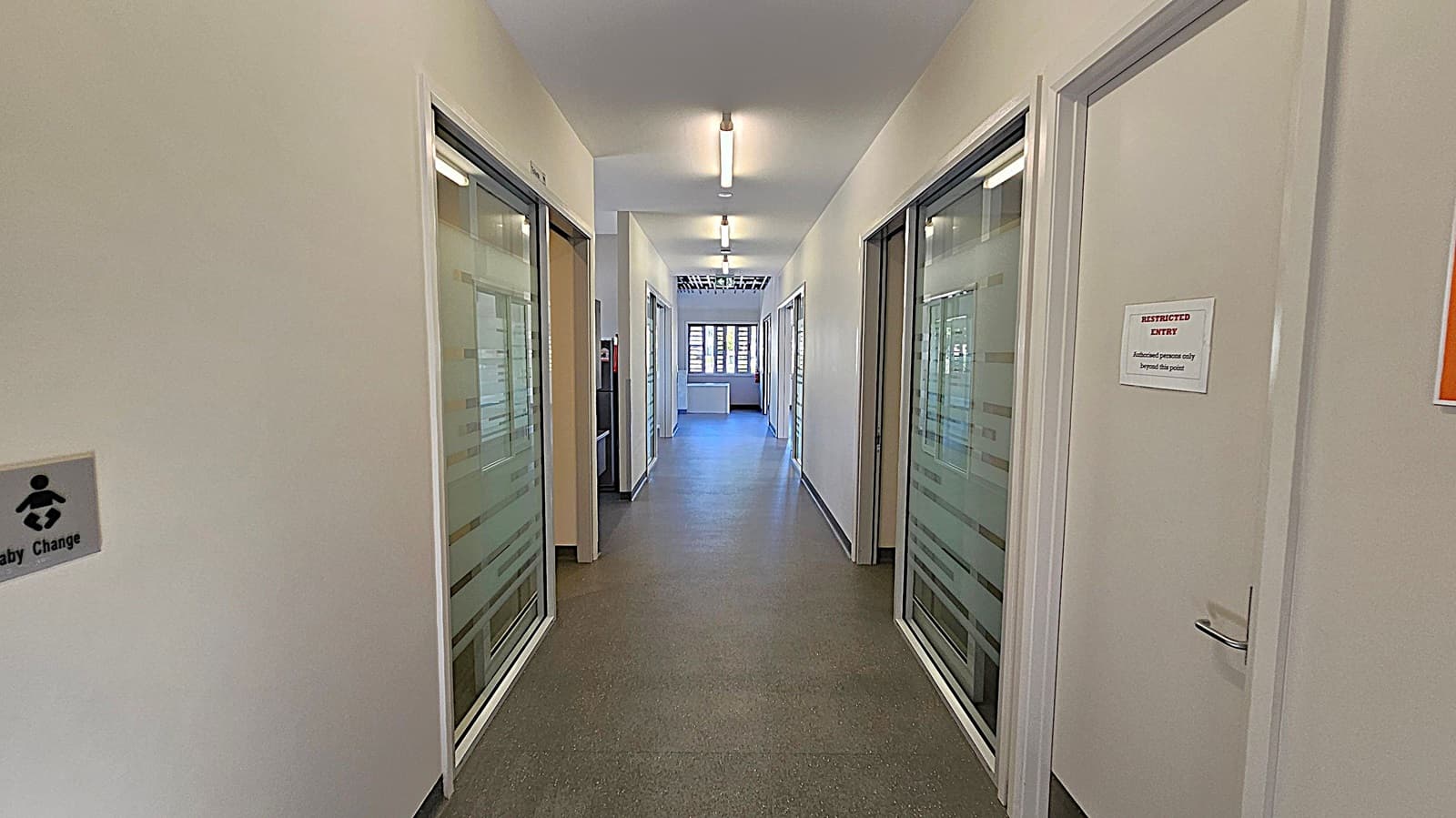 A corridor with a grey speckled floor and white walls. On the left, there's a sign for a baby changing area. On the right, a door with a "RESTRICTED ENTRY" sign. Both sides have glass-panelled doors with frosted stripes. The ceiling has fluorescent lights, and at the end of the corridor, there's a window with horizontal blinds, allowing natural light to enter.