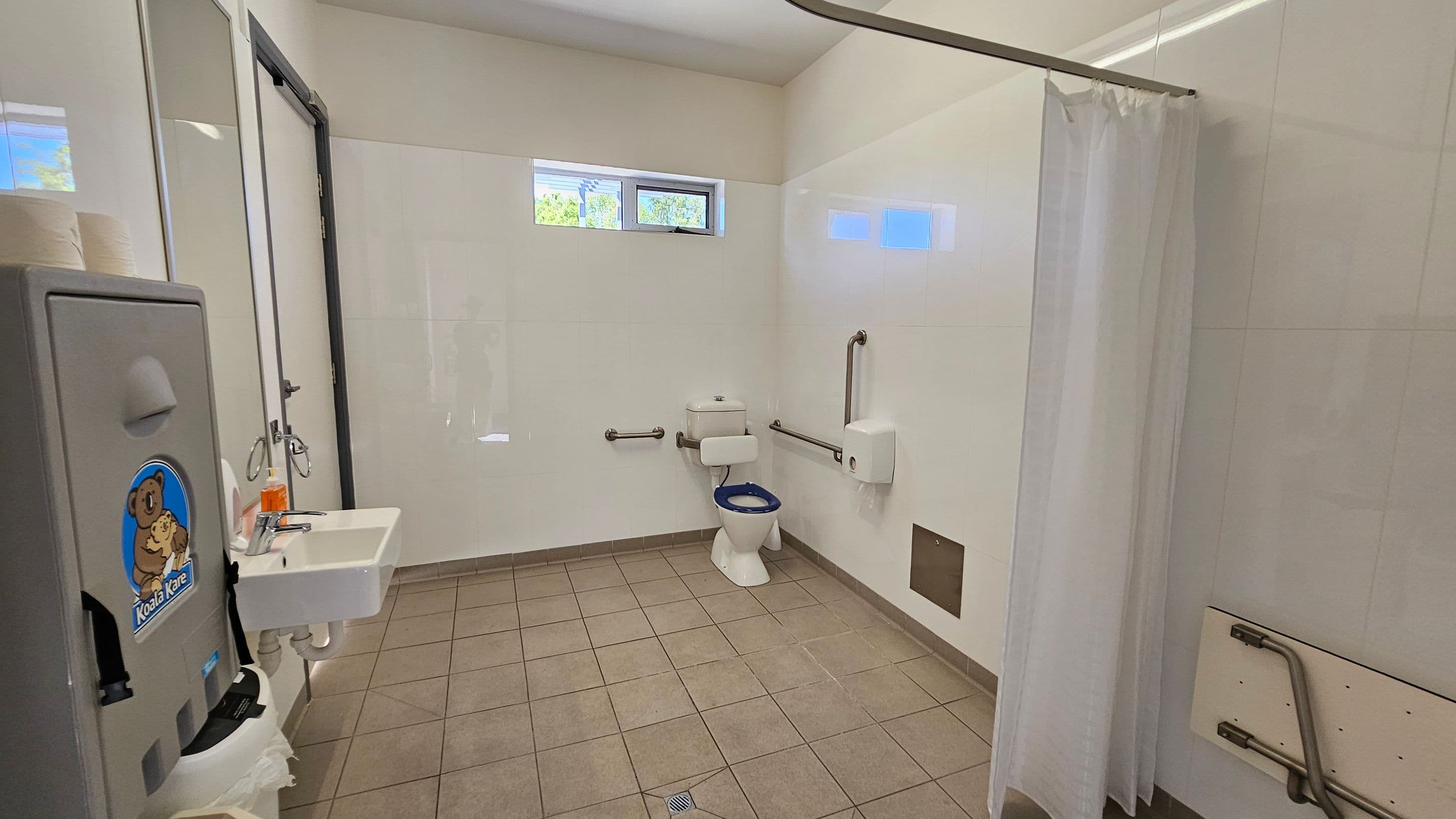A public restroom with a tiled floor and light-coloured walls. On the left, there's a baby changing station with a "Koala Kare" logo, a sink with a soap dispenser, and a mirror. In the centre, a toilet with a blue seat is equipped with grab bars on both sides. On the right, a white shower curtain is partially visible. High windows near the ceiling allow natural light in.
