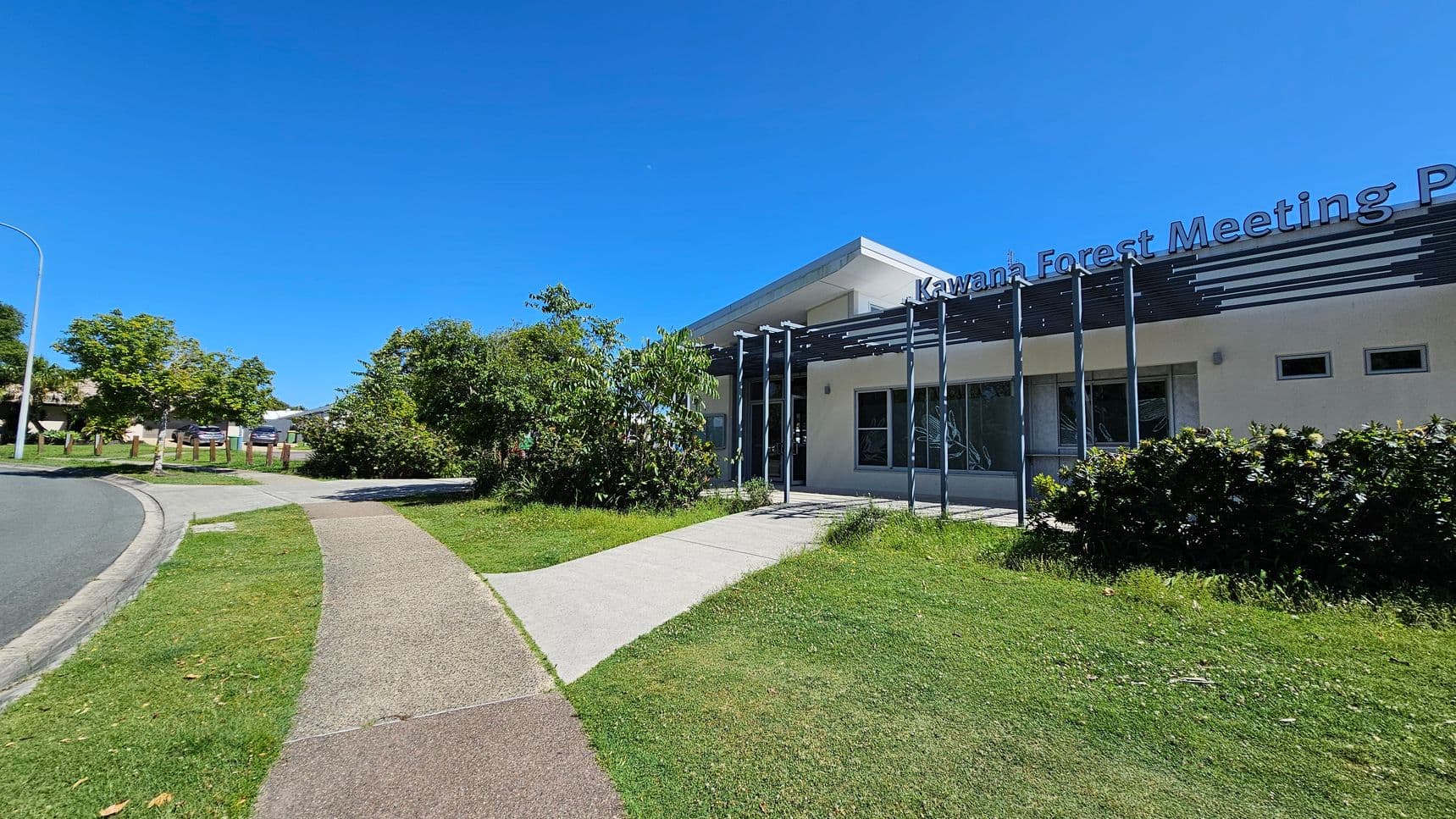 A modern building with a flat roof labelled "Kawana Forest Meeting Place" on the front. The structure has large windows and a pergola with vertical beams. In the foreground, there's a grassy area with a concrete path leading to the entrance. On the left, there are trees and shrubs, while the right side features more greenery and a wooden fence. The sky is clear and blue.