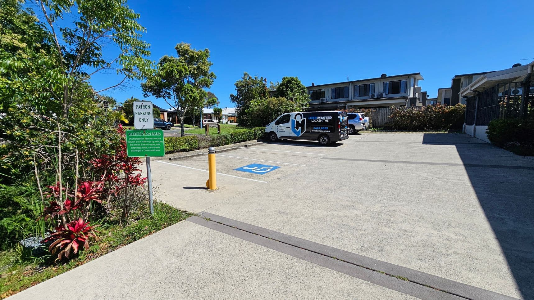 A parking area with a designated disabled parking space marked in blue on the ground. To the left, there are green and red plants with a sign reading "Patron Parking Only" and another about a bioretention basin. A white van with "Lock Master Locksmiths" branding is parked in the background, alongside another vehicle. Trees and shrubs line the perimeter, and a two-storey building is visible in the background. The ground is paved, and a yellow bollard is near the parking space.