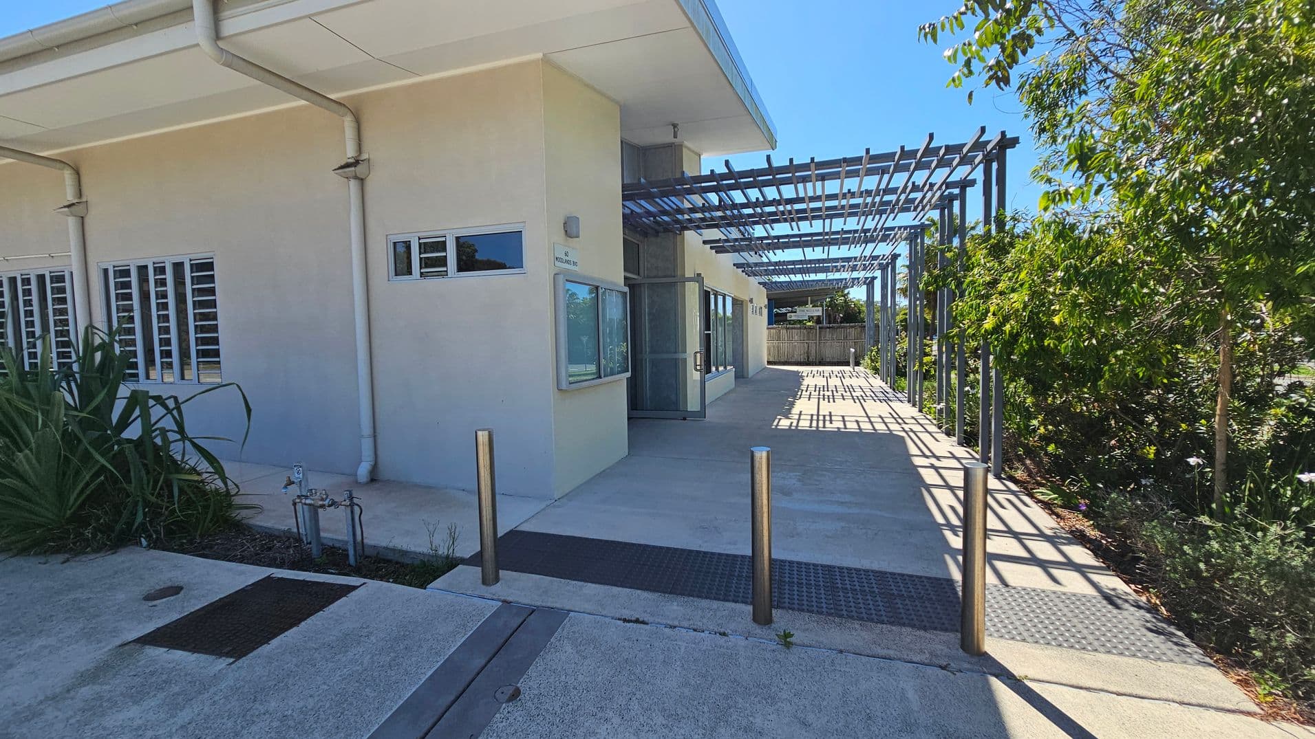 A modern building with a light-coloured exterior and several small windows on the left. The entrance features a glass door and a window with an awning. A metal pergola extends from the building, casting shadows on the concrete path. There are metal bollards near the entrance. On the right, there are trees and shrubs providing greenery. The roof has a slight overhang with visible drainage pipes. The ground is paved with concrete.