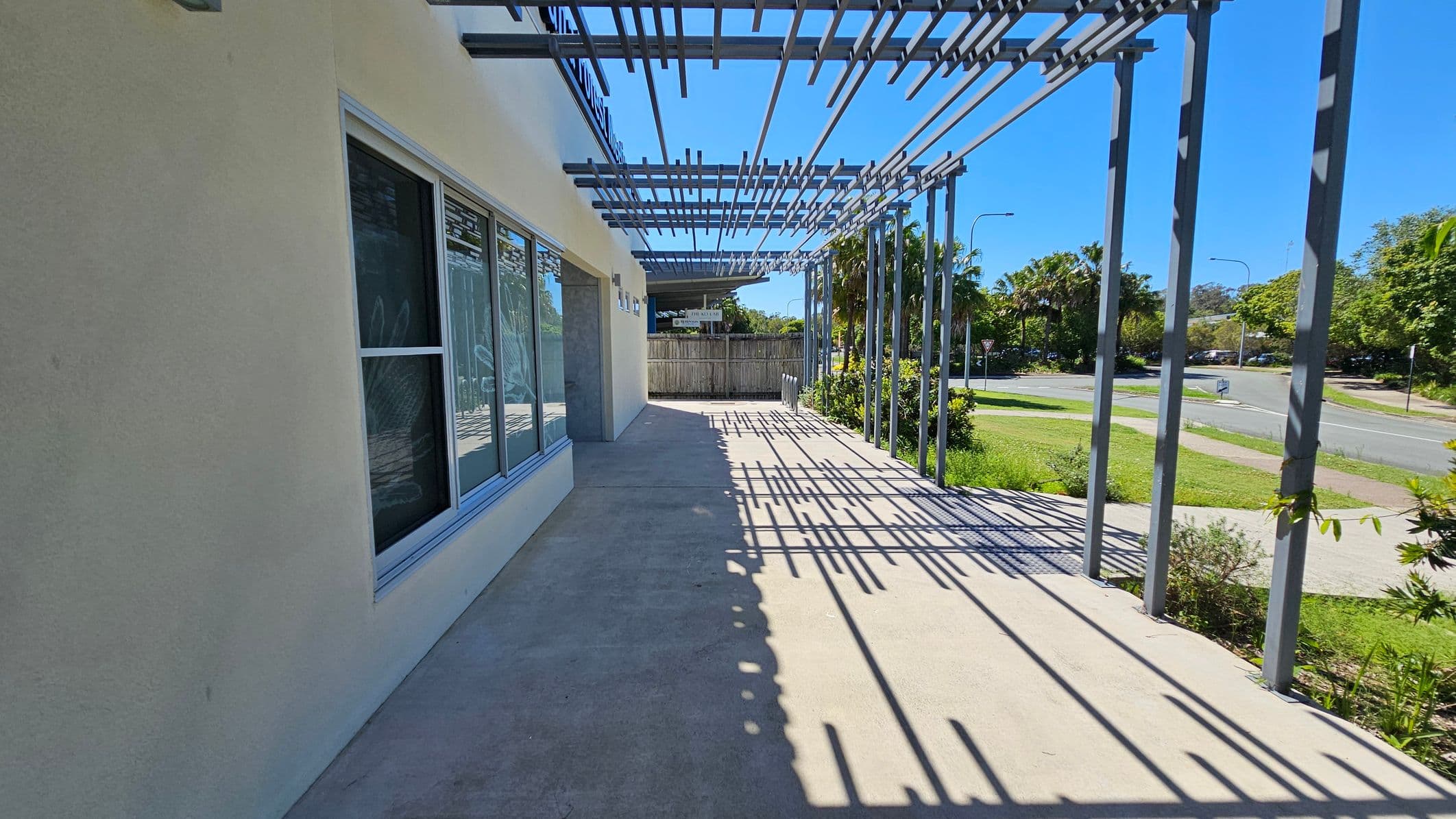 A concrete walkway runs alongside a building with large windows on the left. Overhead, a metal pergola casts intricate shadows on the ground. To the right, a grassy area with bushes and trees lines the street, which curves away in the background. A wooden fence is visible at the end of the walkway.
