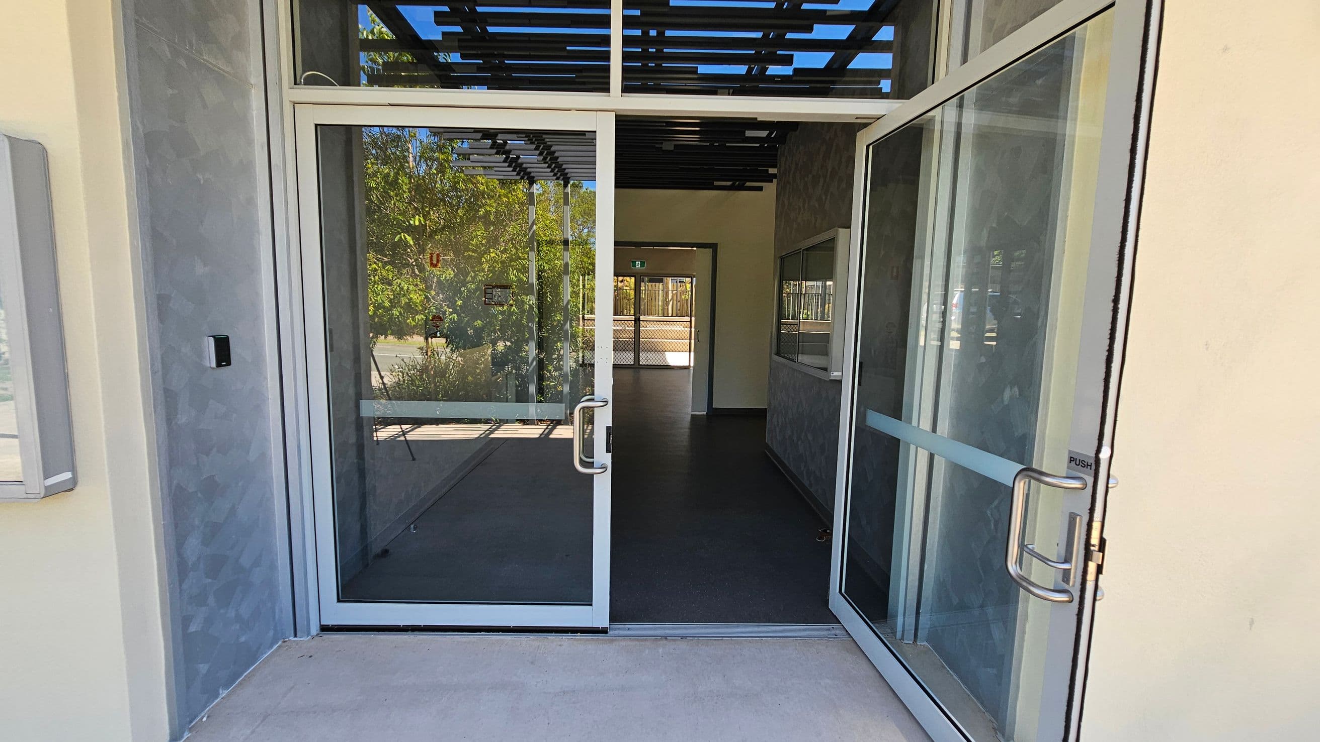 Glass double doors are open, leading into a building. The floor is dark and smooth, and the ceiling has a grid-like structure. Through the doors, there is a view of a corridor with another set of glass doors at the end. Outside, greenery is visible, with trees and plants reflecting on the glass. The walls are light-coloured, and there is a small control panel on the left wall near the entrance.