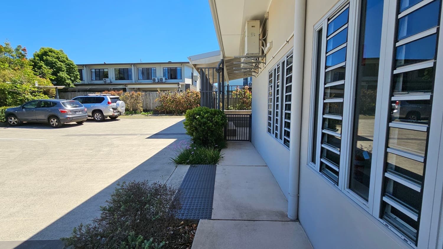 A paved area with a building on the right featuring large windows with horizontal slats. In the foreground, there are small bushes and plants along the path. On the left, a parking area with two cars, a grey and a white one, is visible. In the background, a two-storey building with air conditioning units on the upper level and a wooden fence surrounded by greenery. The sky is clear and blue.