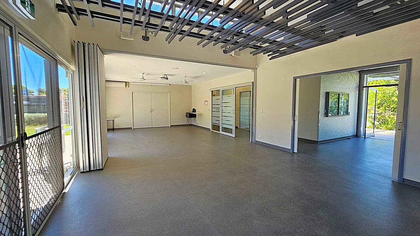 A spacious room with large glass doors on the left and right, allowing natural light to enter. The floor is smooth and grey, while the ceiling features black linear elements and ceiling fans. The background shows a fenced outdoor area with greenery. There are small tables near the walls, and a set of double doors is visible on the right wall.