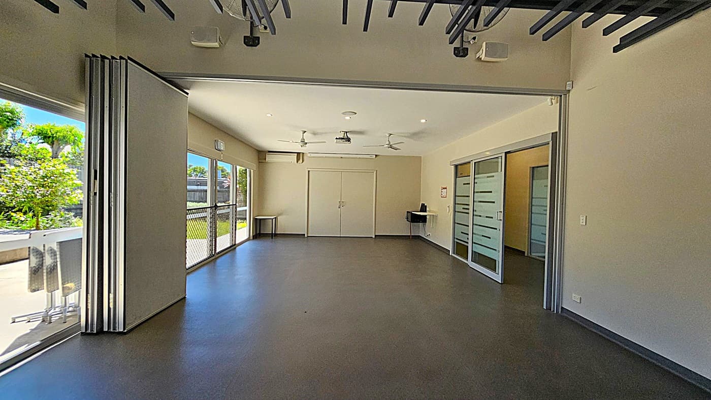 A spacious room with a grey floor and light-coloured walls. On the left, large glass doors open to a view of greenery outside. In the centre, a pair of closed double doors is flanked by a small table. The right side features sliding glass doors with frosted horizontal stripes, leading to another room. The ceiling has exposed beams and is equipped with ceiling fans and recessed lighting.