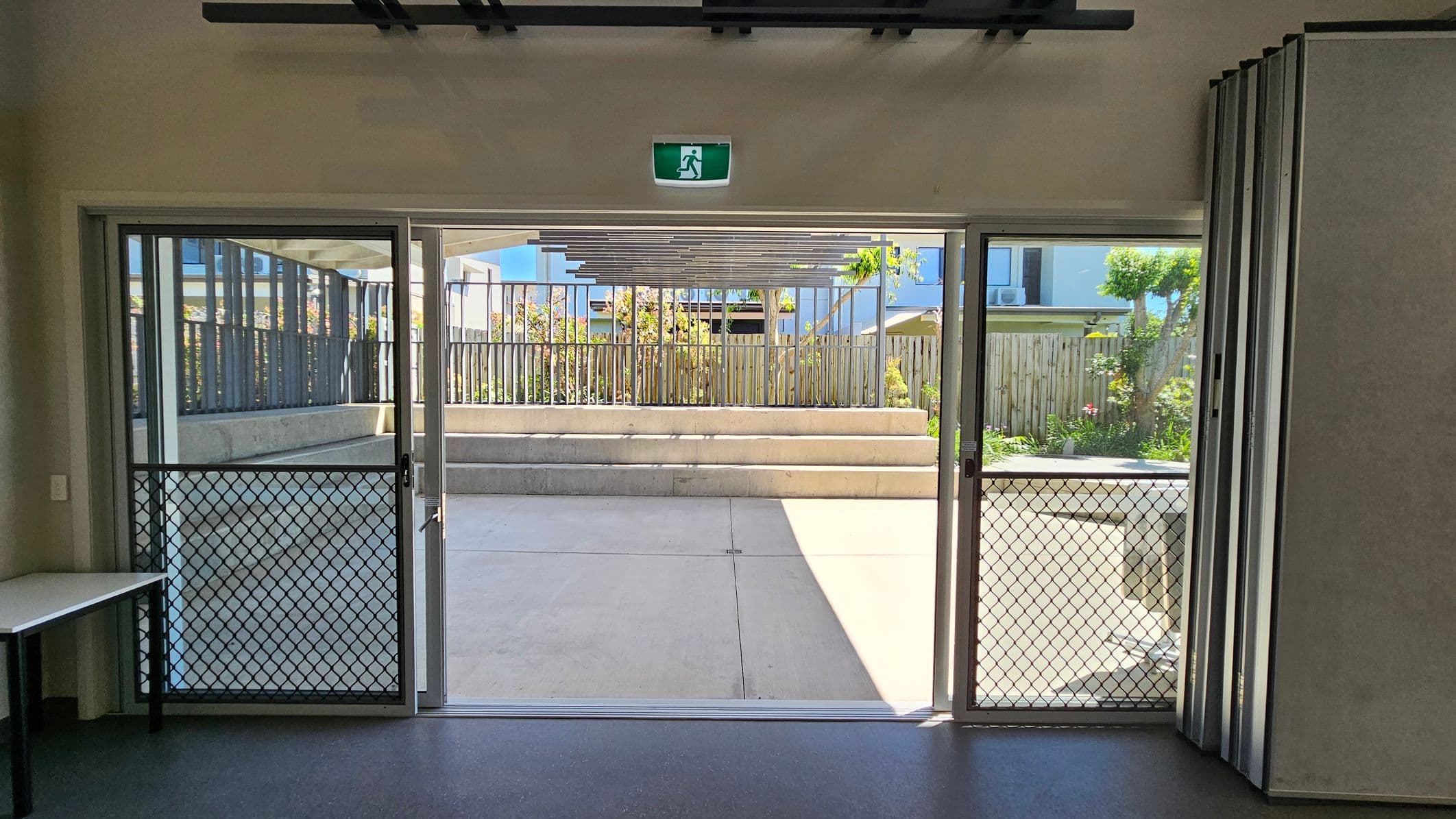 A room with a sliding glass door featuring a mesh screen, leading to an outdoor area. The door is flanked by a small table on the left. Outside, there are concrete steps and a wooden fence with plants and trees visible. Above the door is an illuminated green exit sign. The floor inside is dark, and the ceiling is not visible.