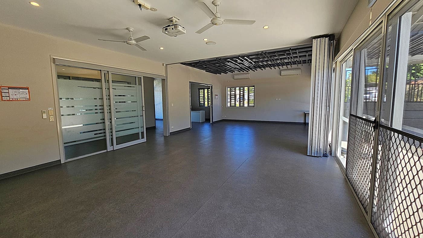 A spacious room with dark flooring and a light ceiling featuring ceiling fans and a projector. On the left, there are frosted glass sliding doors with horizontal stripes. The back wall has a window with horizontal slats and an open doorway leading to another room. On the right, large glass doors with a mesh screen open to the outside. The ceiling has a section with a black grid design.