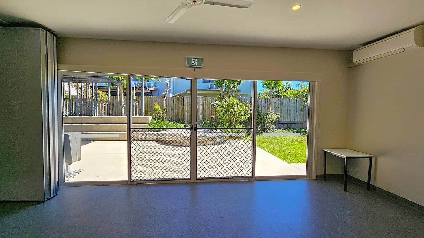 A room with a blue floor and a white ceiling featuring a ceiling fan. On the right, there's a small table and an air conditioning unit mounted on the wall. The back wall has large glass sliding doors with a mesh screen, leading to a garden area with grass, plants, and a wooden fence. An emergency exit sign is above the doors. Outside, there's a concrete path and a circular planter.