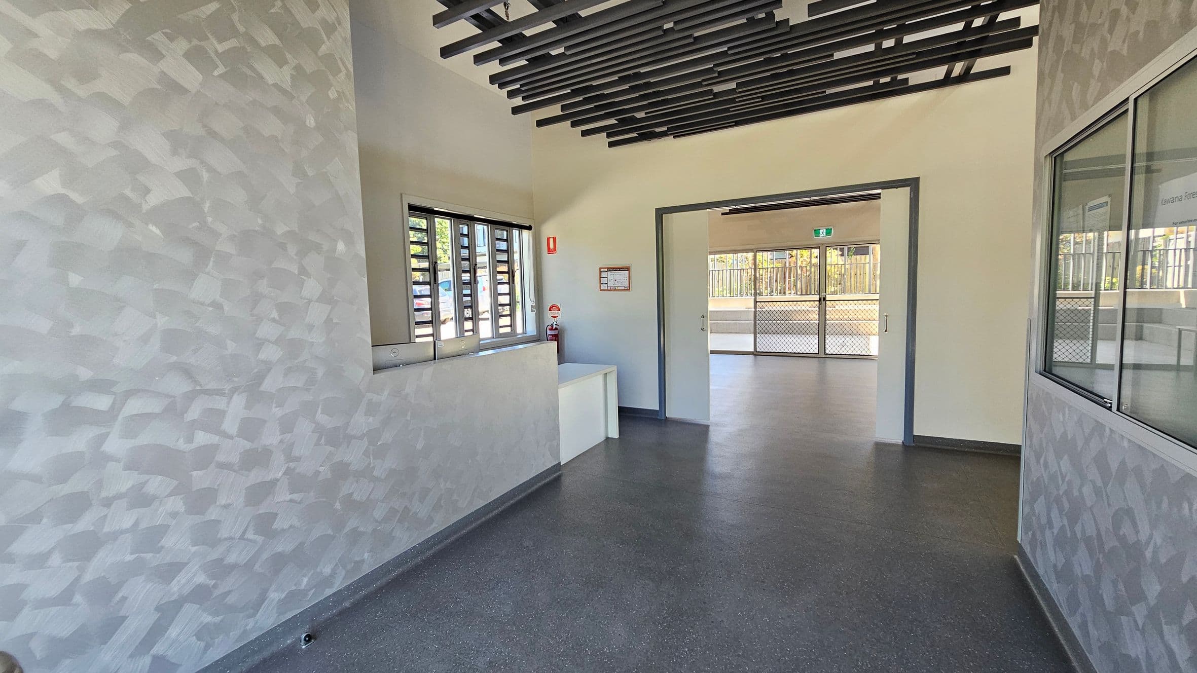 Glass double doors are open, leading into a building. The floor is dark and the walls are light-coloured. A window is on the right wall inside. Outside, there are trees and greenery visible through the glass. The ceiling has a grid-like structure with shadows cast on the ground.