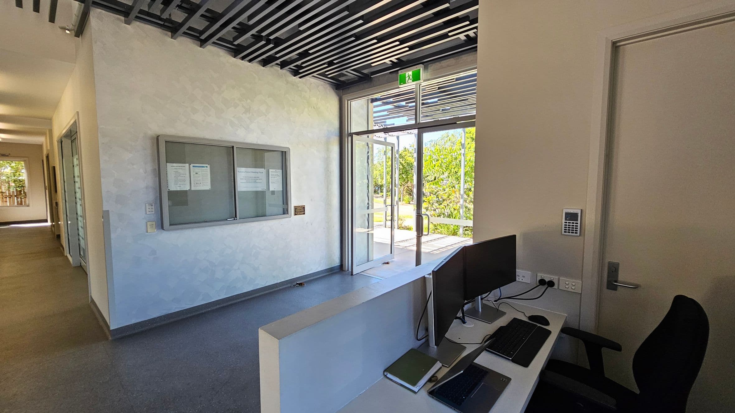 A modern office reception area with a desk on the right, featuring a computer monitor, keyboard, and chair. A door with a keypad is next to the desk. On the left, a corridor leads to other rooms. A notice board is mounted on the wall. The entrance with glass doors is in the centre, showing greenery outside. The ceiling has a grid-like design, and the floor is covered with grey tiles.