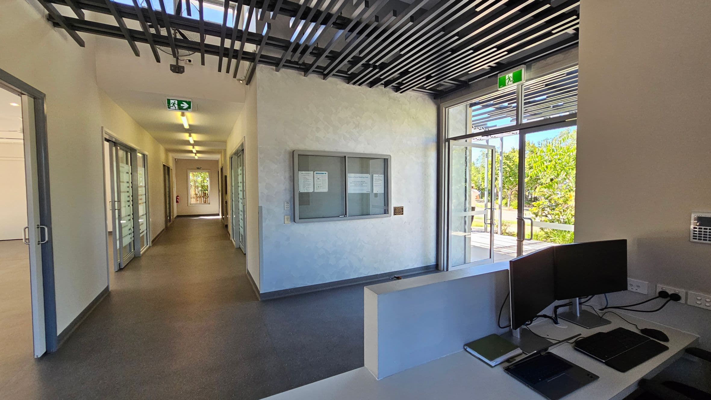 A modern office hallway with a grey carpeted floor and white walls. On the left, there are glass doors leading to offices. In the middle, a notice board is mounted on the wall. On the right, a reception desk with two computer monitors and some electronic devices. The ceiling features a grid-like design. At the end of the hallway, a glass door opens to a garden area with greenery visible outside. An exit sign is above the door.