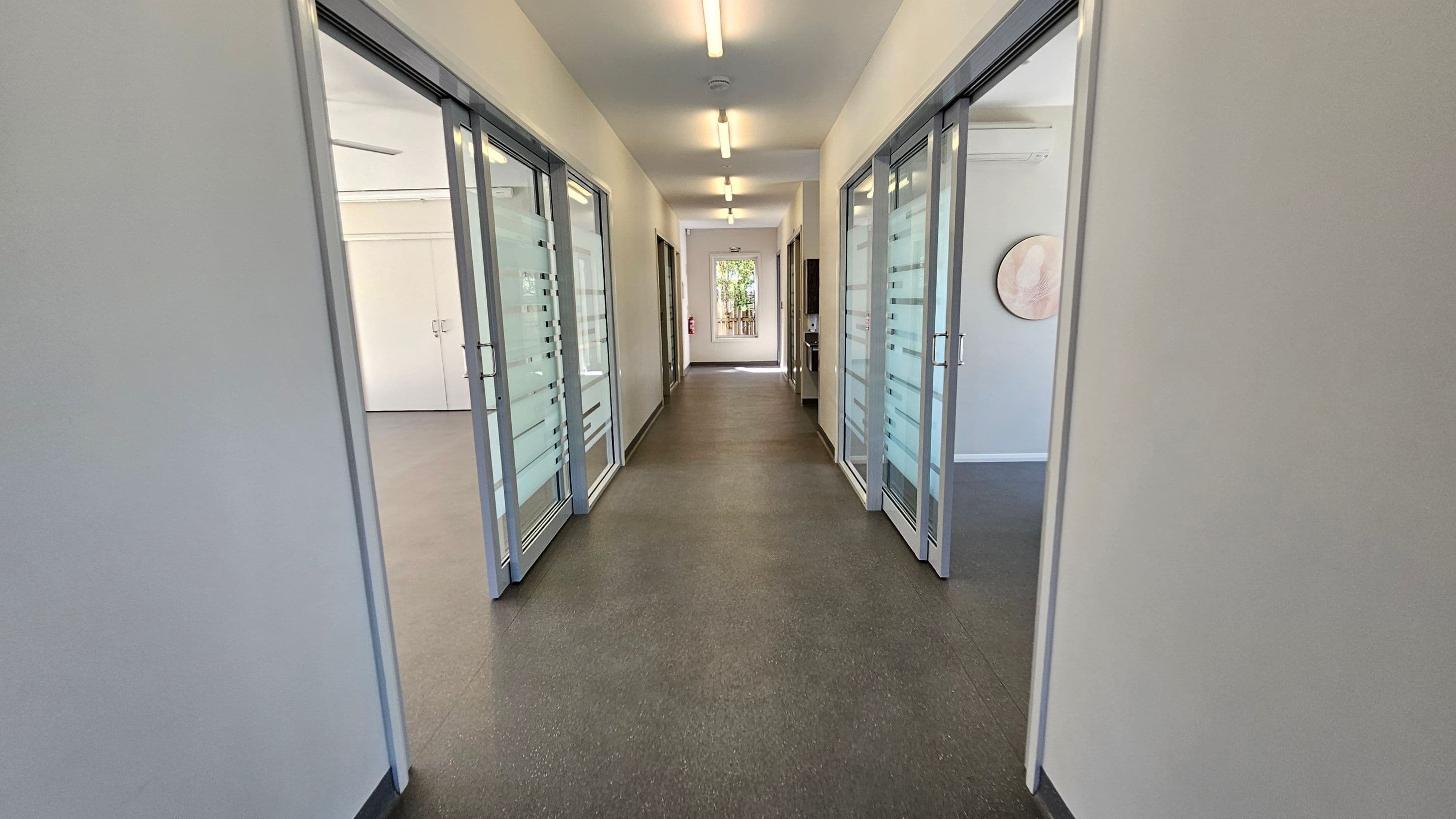 A corridor with a grey and green patterned floor and white ceiling with fluorescent lights. On both sides, there are glass doors with frosted horizontal stripes leading to rooms. At the end of the corridor, a window shows greenery outside. A fire extinguisher is mounted on the left wall near the window.
