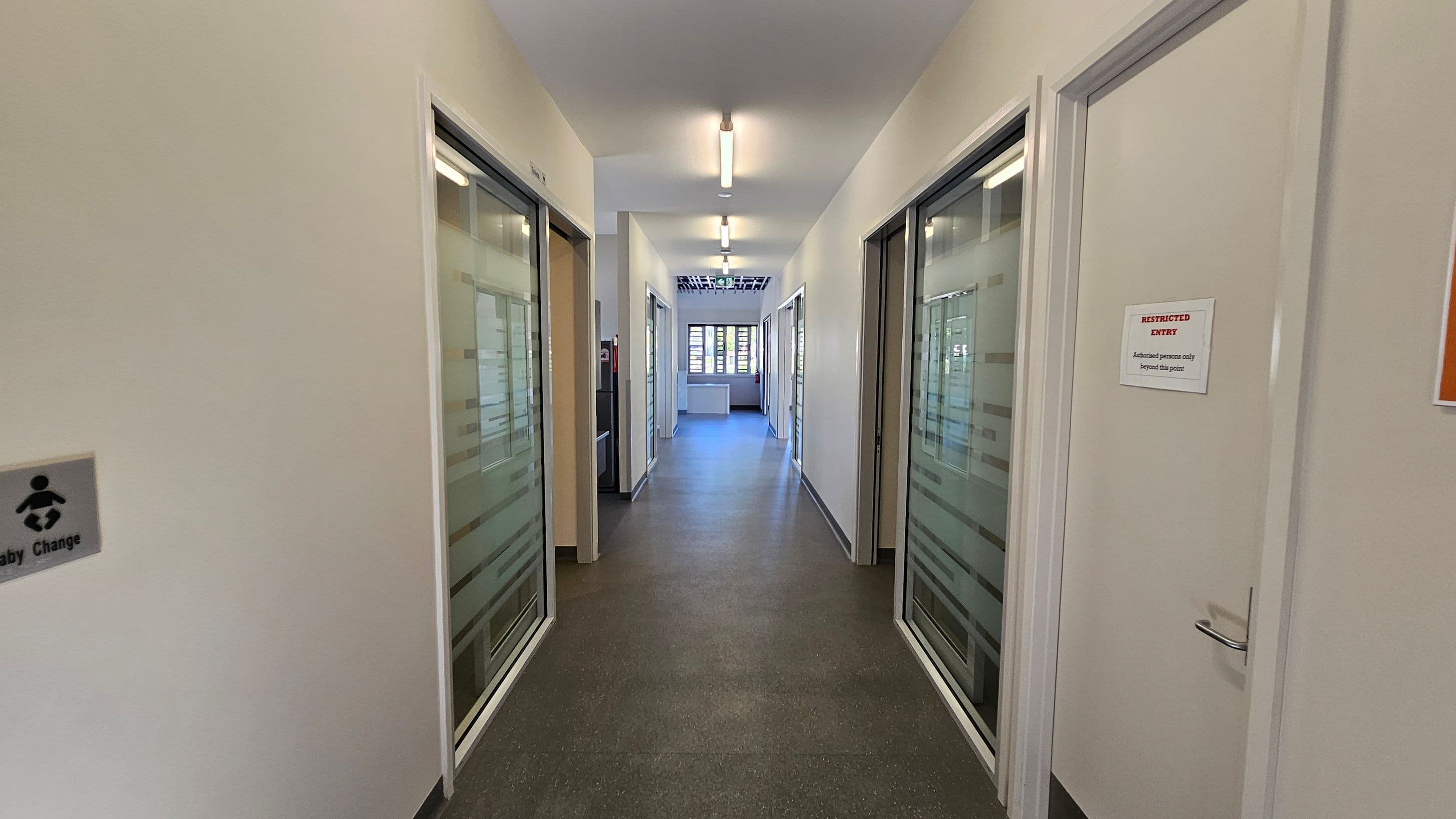A corridor with a grey speckled floor and white walls. On the left, there's a sign for a baby changing area. On the right, a door with a "Restricted Entry" sign. Both sides have glass-panelled doors with frosted stripes. The ceiling has fluorescent lights, and at the end of the corridor, there's a window with horizontal blinds letting in natural light.