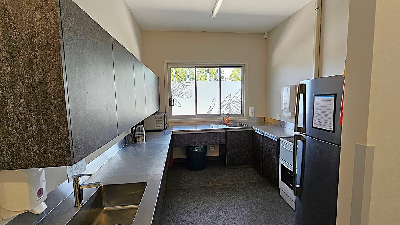A small kitchen with dark wood cabinets and a stainless steel countertop. On the left, there's a sink with a soap dispenser above it. A kettle and microwave are placed on the counter. The back wall features a large window with frosted glass and a view of greenery. Below the window, there's another sink and a bin. The floor is dark grey, and the ceiling has a fluorescent light fixture.