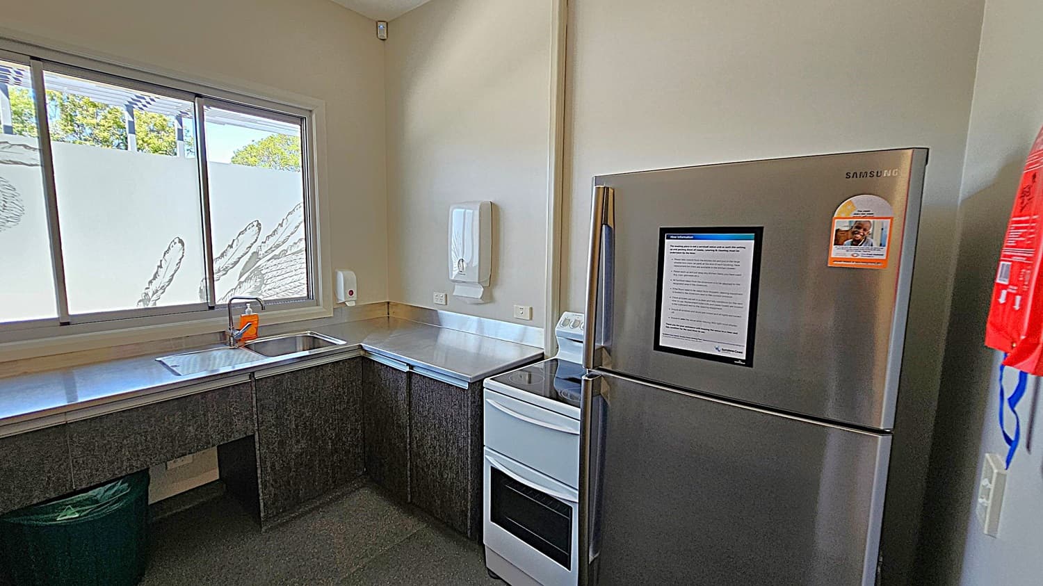 A kitchen with a stainless steel fridge on the right, featuring a notice and a sticker. Next to it is a white oven. On the left, a stainless steel countertop with a sink and a window above it, partially covered with frosted glass and decorative patterns. A soap dispenser and paper towel holder are mounted on the wall. The floor is dark, and the walls are light-coloured.