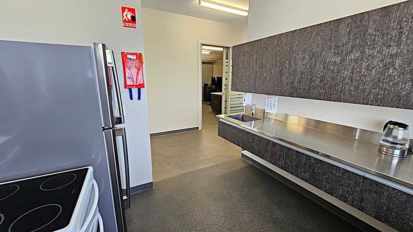 A kitchen area with a grey refrigerator and an electric stove on the left. A fire blanket is mounted on the wall above. On the right, there is a long stainless steel countertop with a sink and a kettle. Dark textured cabinets are above the counter. The floor is a speckled grey, and the ceiling has fluorescent lighting. An open doorway leads to another room with office furniture visible in the background.