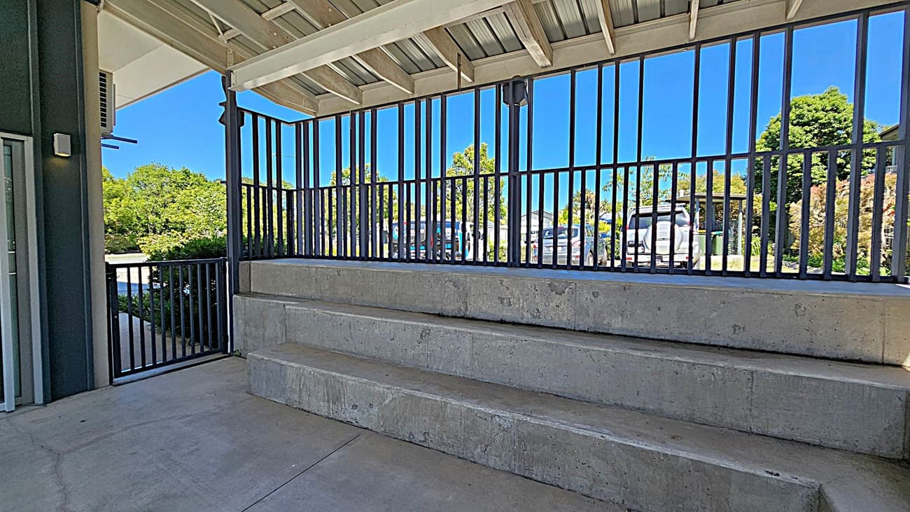Concrete seating steps are arranged in a U-shape around a paved area. Metal railings line the perimeter, with a view of parked cars and trees outside. The ceiling features a slatted design with beams, allowing light to filter through. The floor is smooth concrete, and the space is partially shaded.