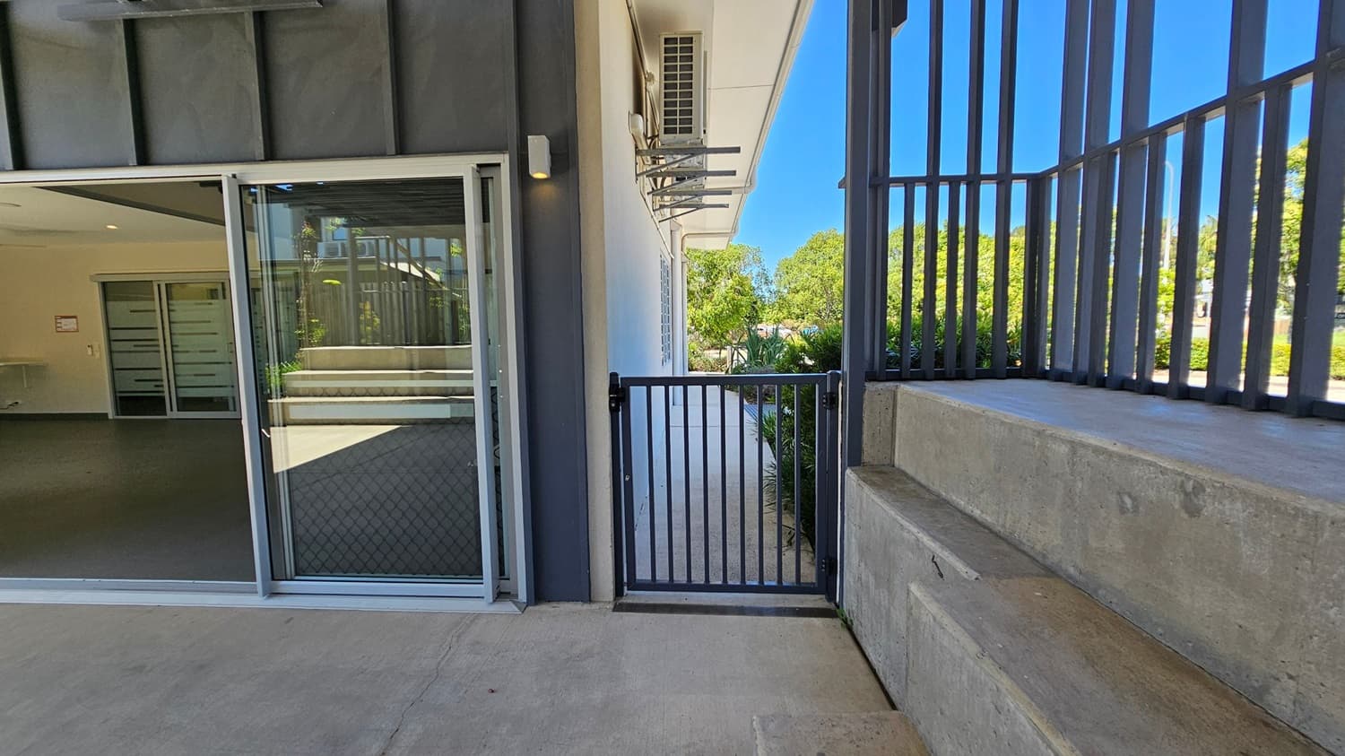 A concrete pathway leads to a metal gate in the centre, with a building on the left featuring a glass sliding door and a visible air conditioning unit above. On the right, there is a concrete wall with metal railings. In the background, greenery and trees are visible under a clear blue sky.