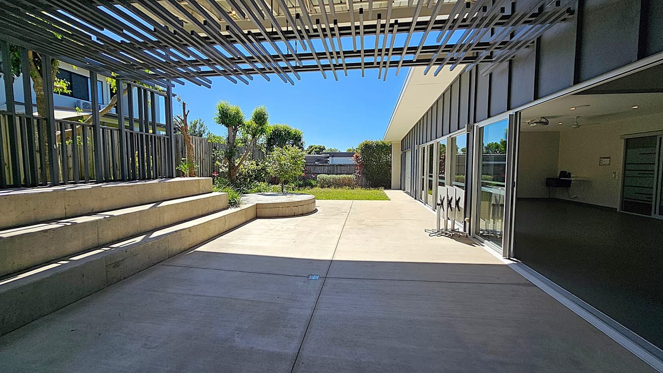 A modern outdoor space with a concrete floor and a slatted pergola overhead casting shadows. On the left, there are tiered concrete steps and a wooden fence with greenery. In the centre, a circular concrete planter with plants and a grassy area. On the right, a building with large glass doors, some open, revealing an interior space. A bike rack is visible near the doors. The background features more greenery and a wooden fence.