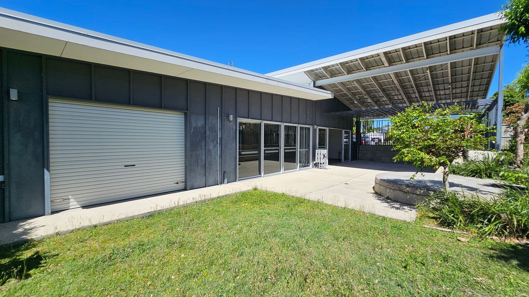 A modern building with a grey exterior and a large roller door on the left. To the right, there are glass sliding doors and a covered area with a slanted roof. A small tree and some greenery are in front of the building, with a concrete path leading to the entrance. The ground is covered with grass, and the sky is clear.