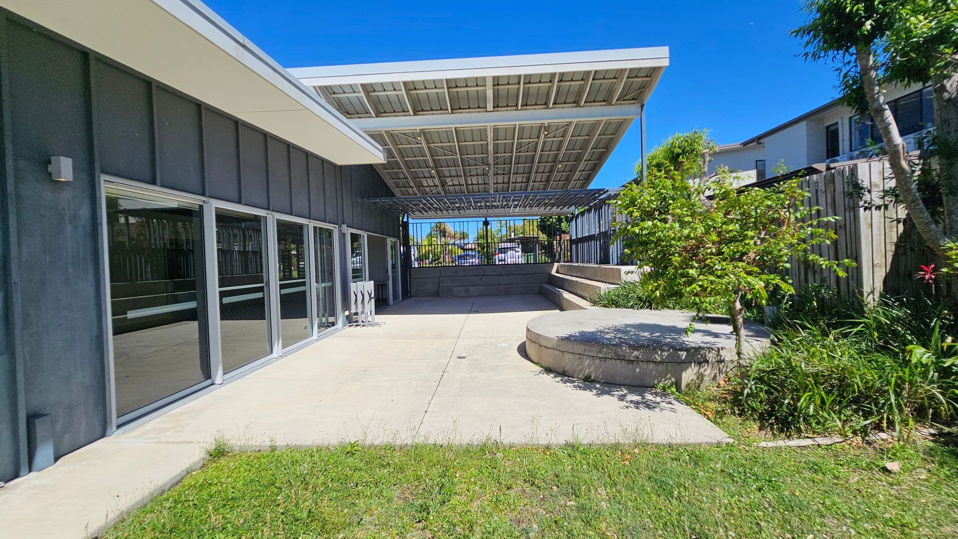 A modern outdoor area with a concrete patio and a grassy foreground. On the left, there is a building with large glass doors and a dark exterior. In the centre, a large metal canopy provides shade. To the right, there are raised concrete seating areas surrounded by greenery, including small trees and plants. A wooden fence and neighbouring buildings are visible in the background.