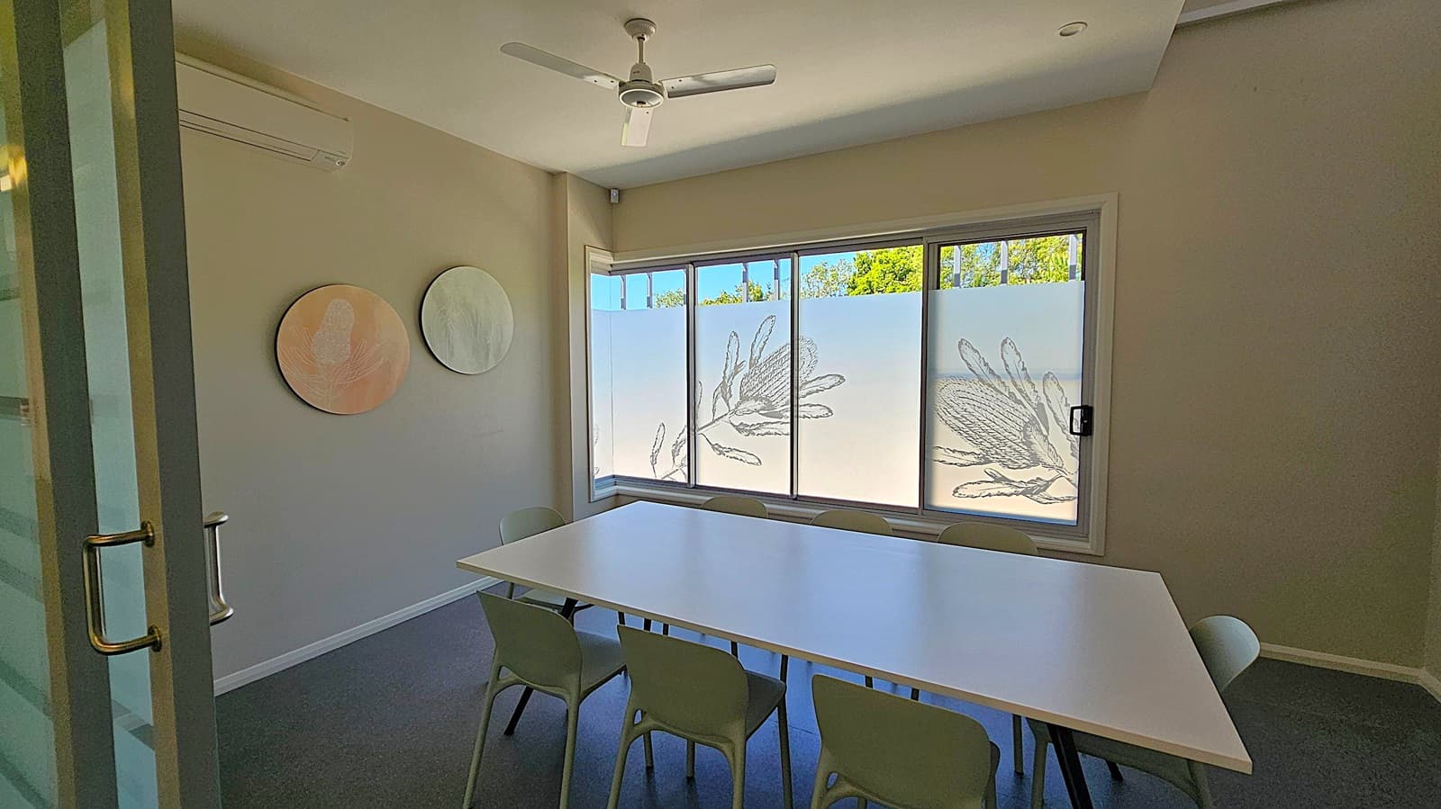 A small meeting room with a white rectangular table surrounded by six chairs. On the right wall, a flat-screen TV is mounted. The left side shows a glass door with a metal handle. The back wall features large windows with frosted glass and decorative plant designs, allowing some natural light. The ceiling has a white fan, and the floor is carpeted in a dark colour.