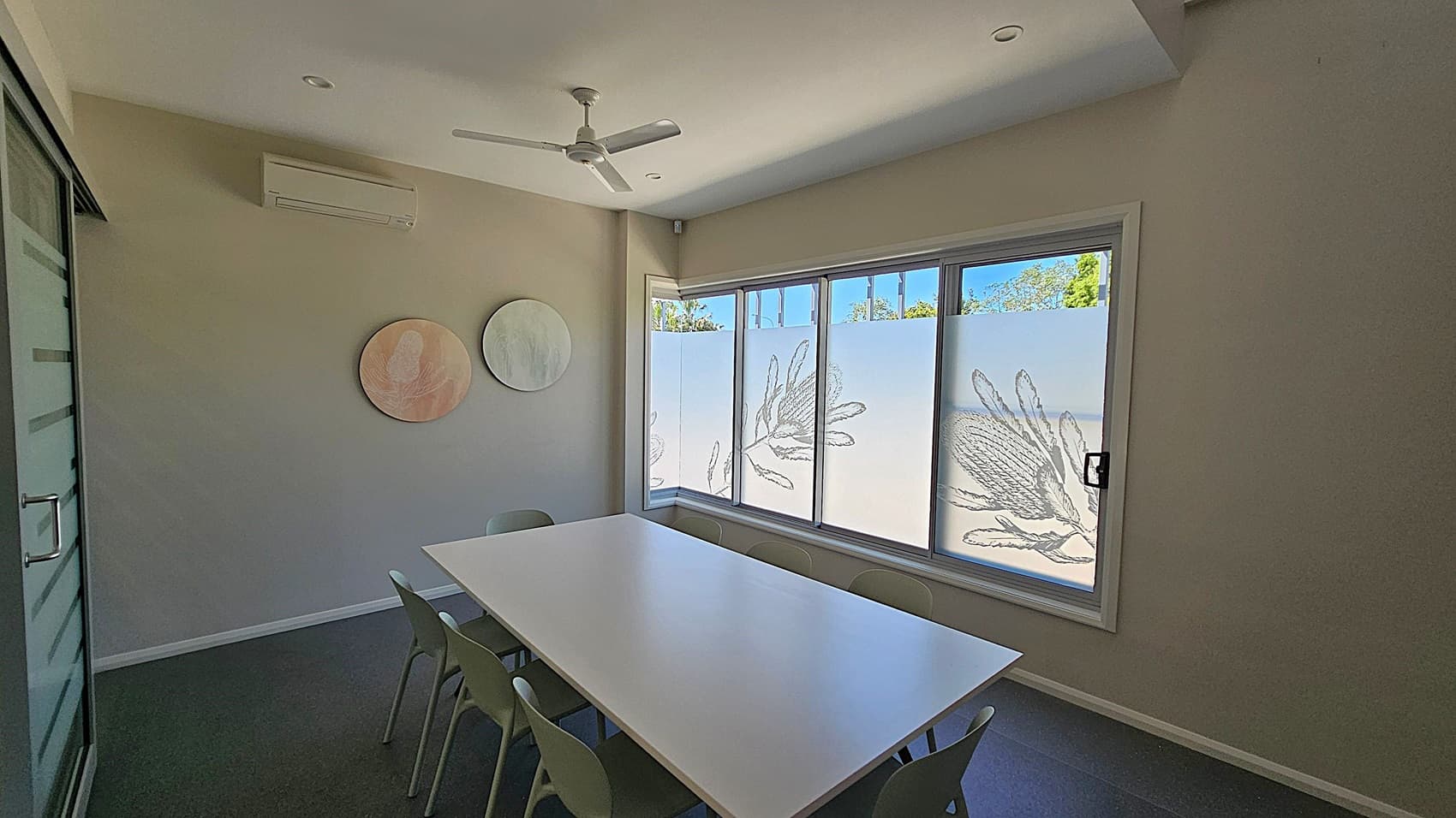 A small meeting room with a long white table surrounded by green chairs. On the left wall, there are two circular artworks. The right side features large windows with frosted glass, decorated with botanical designs. An air conditioning unit is mounted on the left wall above the artworks. The ceiling is white with recessed lighting and a ceiling fan. The floor is dark-coloured.