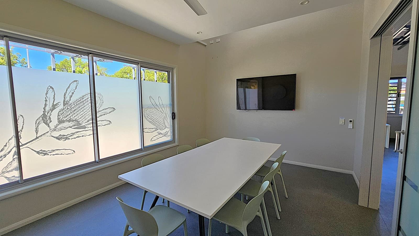 A small meeting room with a long white table surrounded by eight chairs. On the right wall, a flat-screen TV is mounted. The left side features large windows with frosted glass, decorated with floral designs, allowing some natural light. The floor is dark grey, and the ceiling is light-coloured with recessed lighting. A partially open door on the right leads to another room.