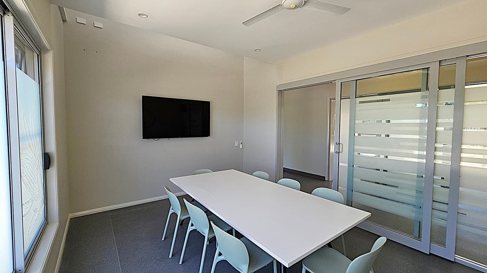 A small meeting room with a rectangular white table surrounded by eight light blue chairs. On the left wall, there's a large window. A flat-screen TV is mounted on the wall opposite the window. The right side features sliding glass doors with frosted horizontal stripes. The ceiling has a white fan and recessed lighting. The floor is dark grey.