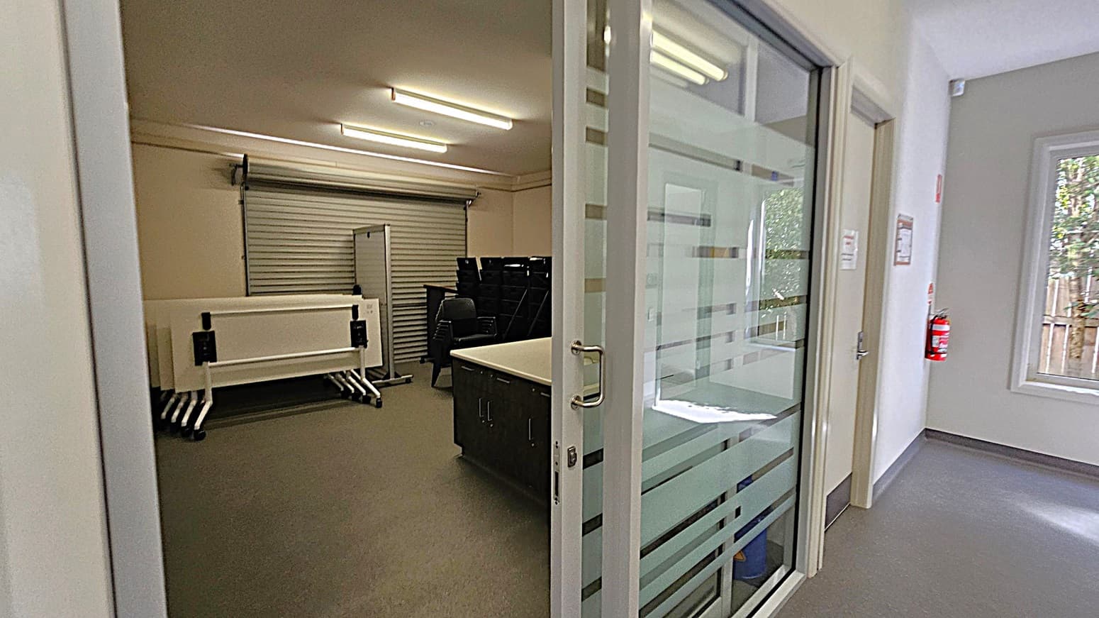 A room with a glass sliding door on the right, featuring horizontal frosted stripes. Inside, there are stacked tables and chairs against a wall with a closed roller shutter. The floor is grey, and the ceiling has fluorescent lights. In the hallway, a fire extinguisher is mounted on the wall near a window with a view of trees and a wooden fence outside.