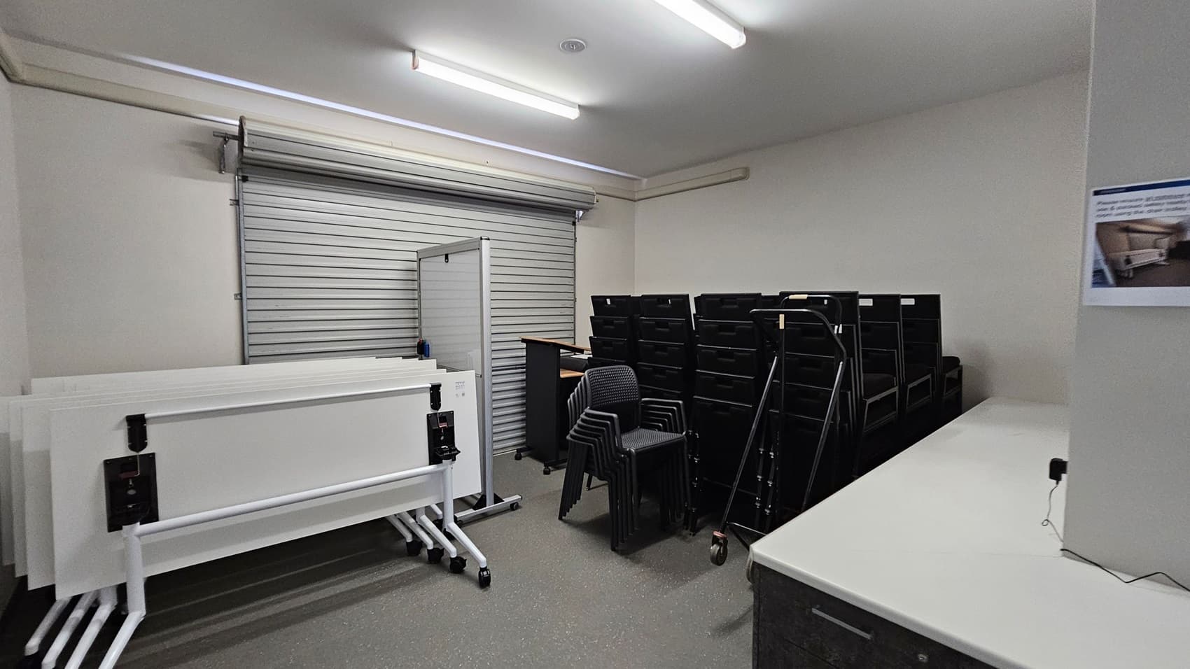A storage room with a grey roller shutter door on the left. In front of it, several white folding tables are stacked on wheeled frames. In the centre, there are stacked black chairs and a trolley. On the right, a row of black storage units is visible. A white countertop runs along the right wall, with a poster above it. The floor is grey and the ceiling has fluorescent lights.