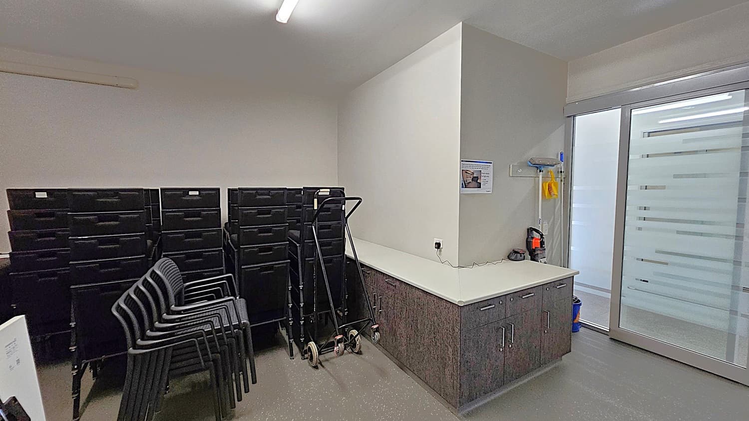 A room with a stack of black chairs and several black storage bins on the left. In the centre, a hand truck is leaning against a white wall. On the right, a countertop with dark cabinets below, a fire extinguisher, and cleaning supplies hanging on the wall. A large glass sliding door with frosted horizontal stripes is on the far right. The floor is grey with a speckled pattern, and the ceiling has a fluorescent light fixture.