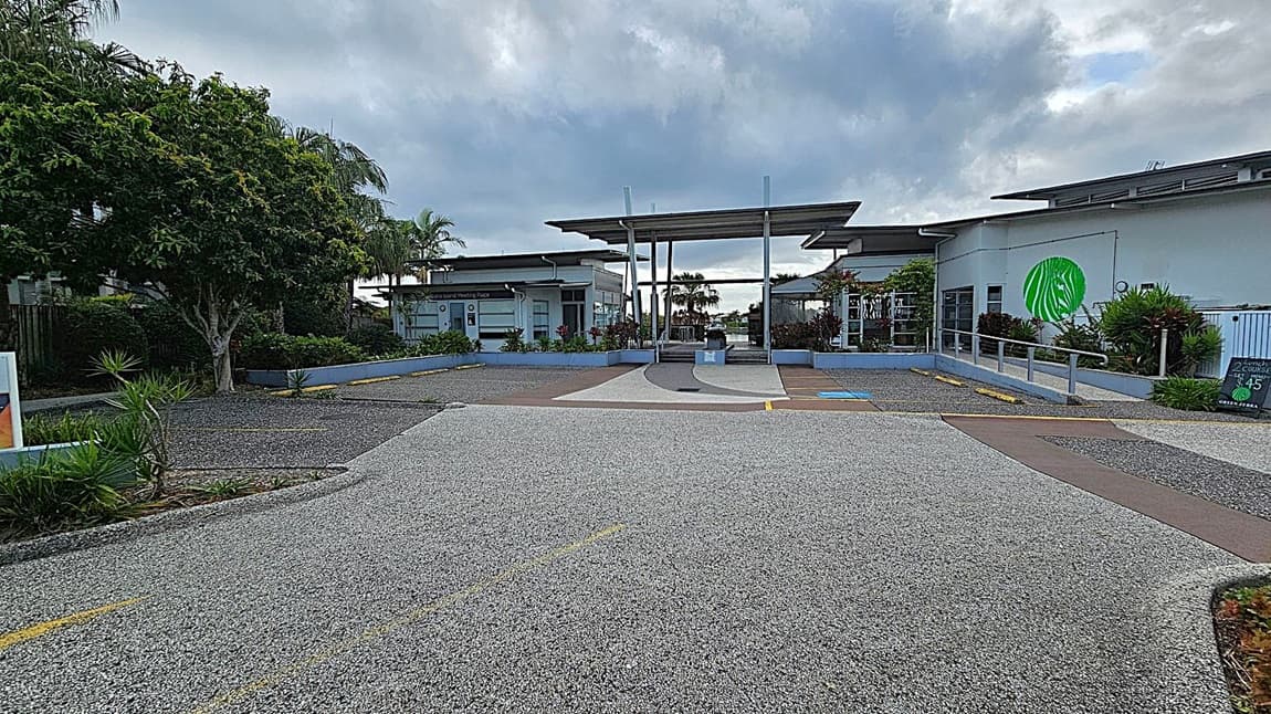 A pathway with yellow directional arrows leads to a modern building with a flat roof. On the left, there are palm trees and a sign with the number 5, indicating "Kawana Island Meeting Place." On the right, a black sign advertises the "Green Zebra Restaurant & Bar" with lunch specials and hours. The area is landscaped with green shrubs and plants. The building features large windows and a logo with a green zebra.