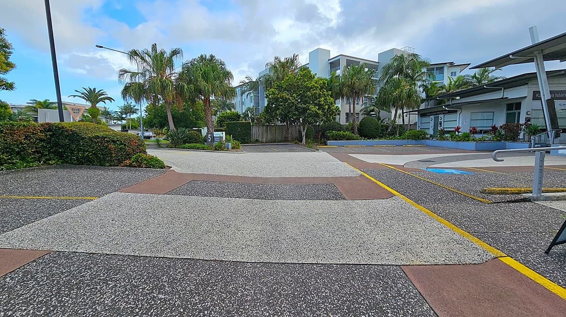 A street scene with a textured, multi-coloured pavement featuring grey, brown, and black sections. On the left, there are lush green bushes and palm trees, with a "Give Way" sign visible. In the centre, a modern white building is partially obscured by more palm trees and greenery. On the right, a building with a sloped roof and a sign is surrounded by landscaped plants. A metal railing is visible in the foreground.